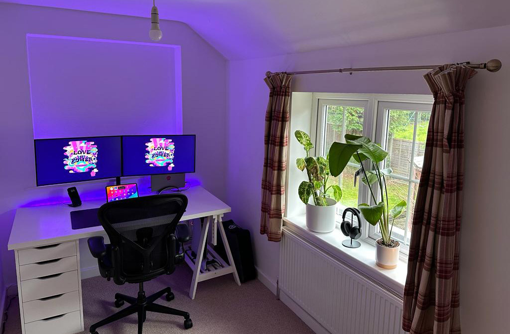 A full view of my new setup. A white desk with drawers on the left, and a trestle leg on the right. A monitor arm holds two monitors in places, and behind the screens and desk there is ambient purple light. To the right, two plants sit on a windowsill, either side of a pair of black headphones on a stand